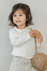 Toddler in Avauma Chloe Ribbon T-shirt in Cotton Cream, holding a woven basket with a joyful smile.